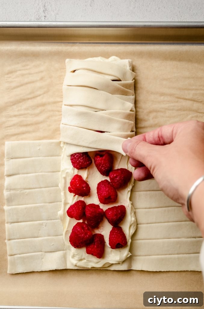 Raspberry Cream Cheese Danish Recipe 11 Someone is folding the strips of a puff pastry filled with cream cheese and raspberries to make a pastry braid.