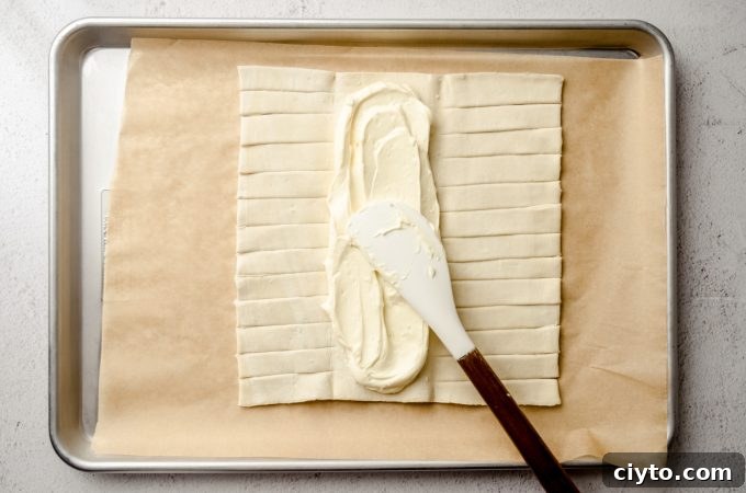 Raspberry Cream Cheese Danish Recipe 8 A rectangle of puff pastry dough on a baking sheet lined with parchment paper and someone is spreading a cream cheese filling into the center flap.