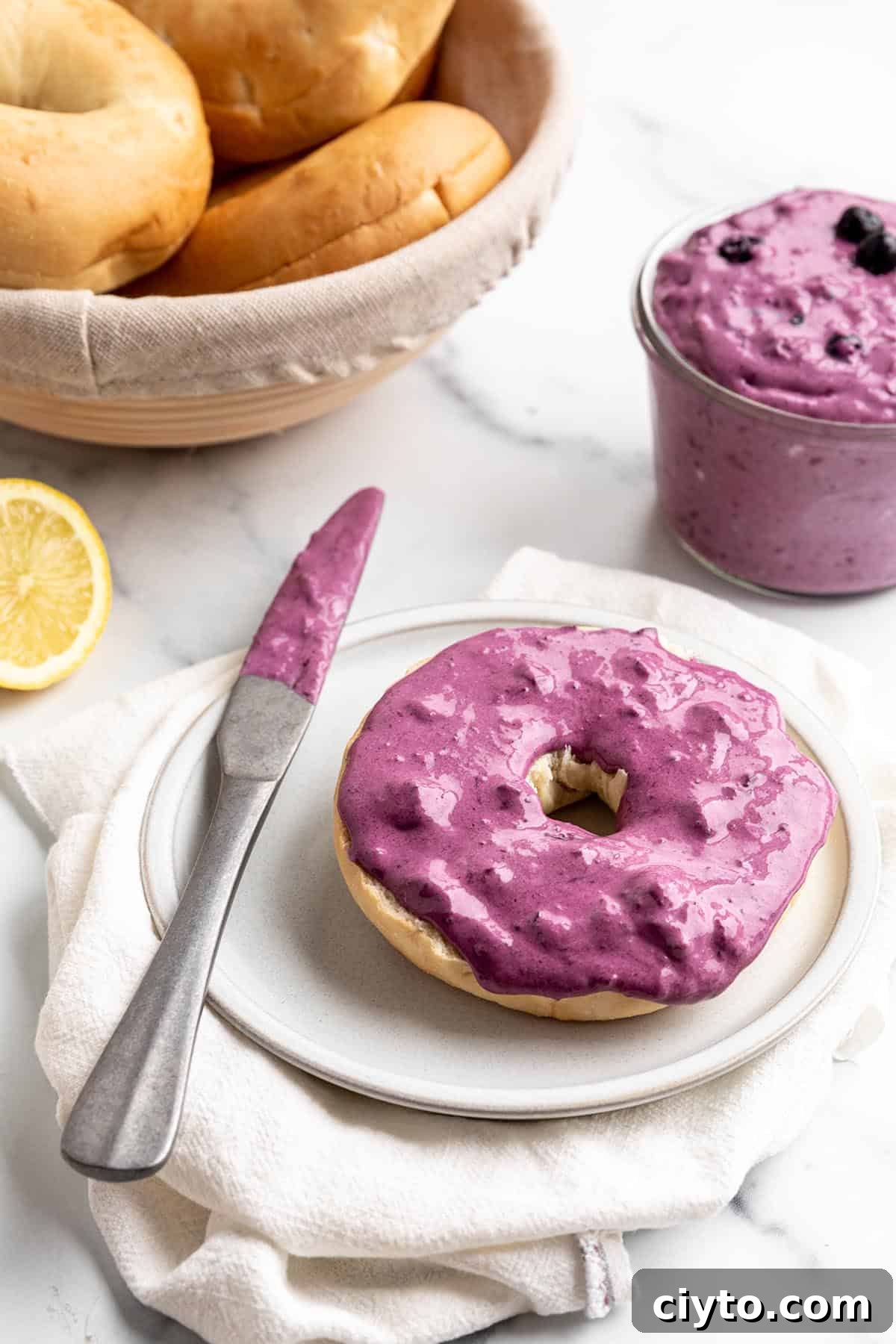 A bagel with a blueberry cream cheese spread, basket of bagels and a fresh lemon.