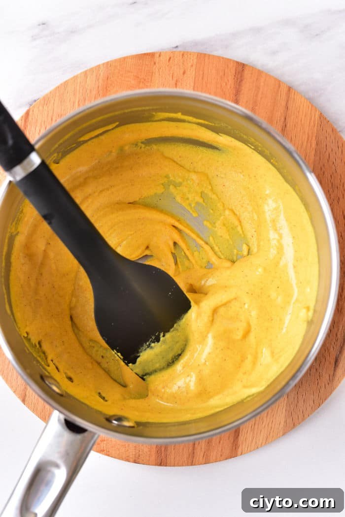 A saucepan filled with creamy hot dog mustard, being stirred by a black spatula on a wooden trivet.