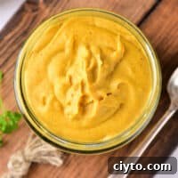 Glass jar of creamy yellow hot dog mustard on a wooden board with a spoon beside it.