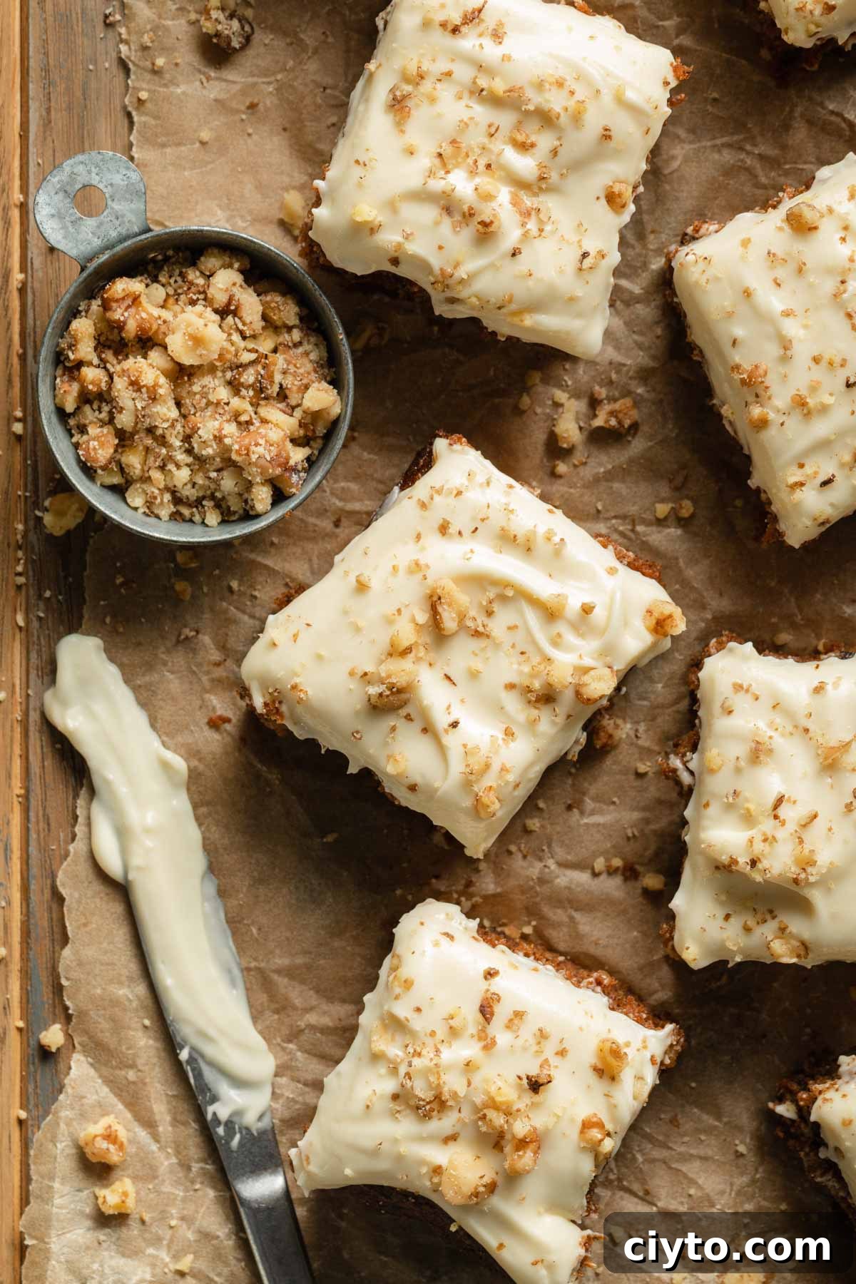 Overhead view of pieces of carrot cake arranged on a sheet of crumpled brown parchment paper next to a cup of chopped walnuts.