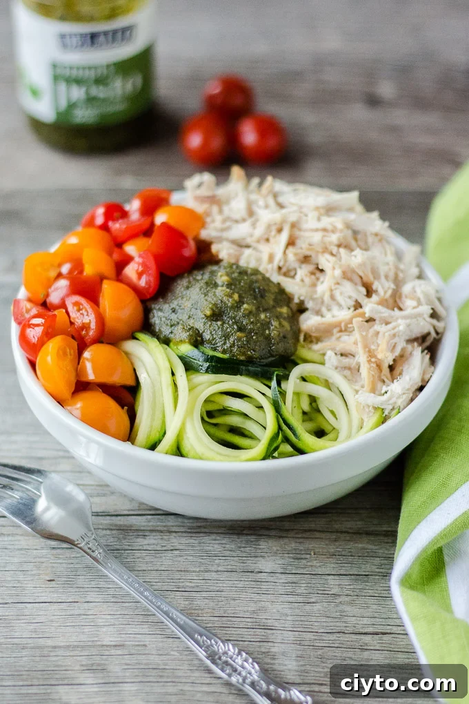 white bowl with pesto, chicken, zoodles, and cut tomatoes on a gray background with a white and green towel on the right, silver fork on the left, and pesto container and 3 cherry tomatoes in the background