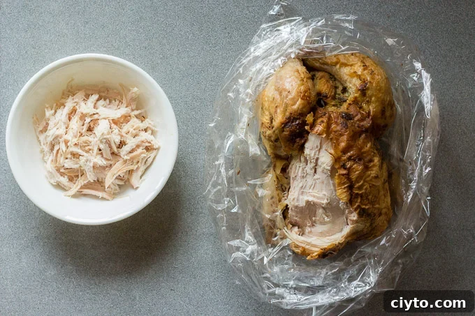 a rotisserie chicken on the right in a plastic bag with the left breast shredded into a white bowl on the left