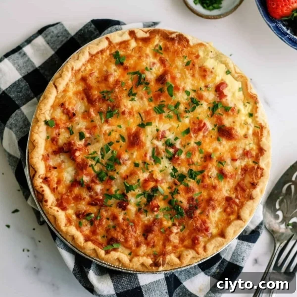 A golden-brown quiche garnished with chopped herbs sits on a checkered cloth. A pie server is beside it, with small bowls of chopped herbs and strawberries nearby.