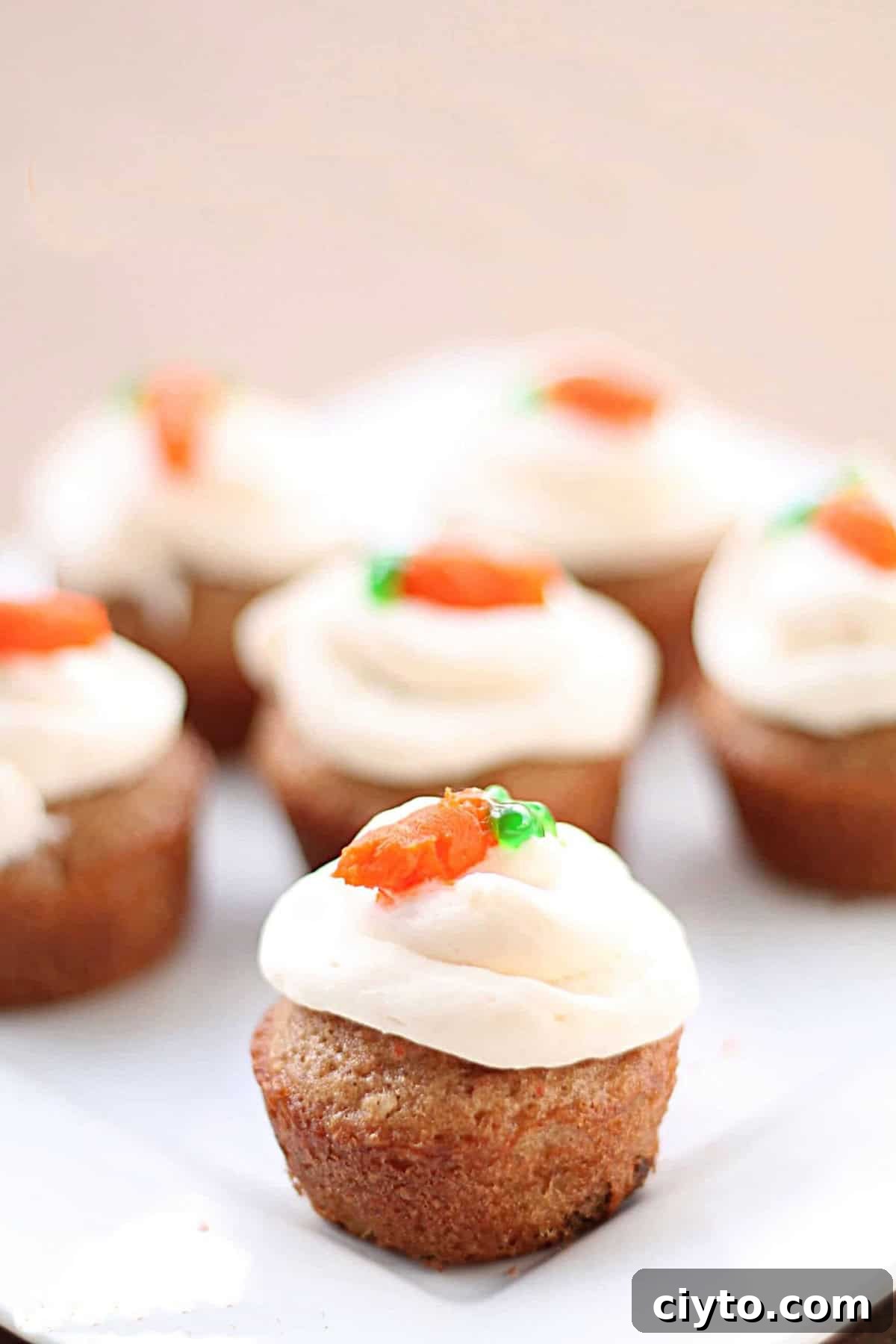 A close-up of mini cupcakes with white frosting and small orange and green decorations on top, arranged on a white plate.