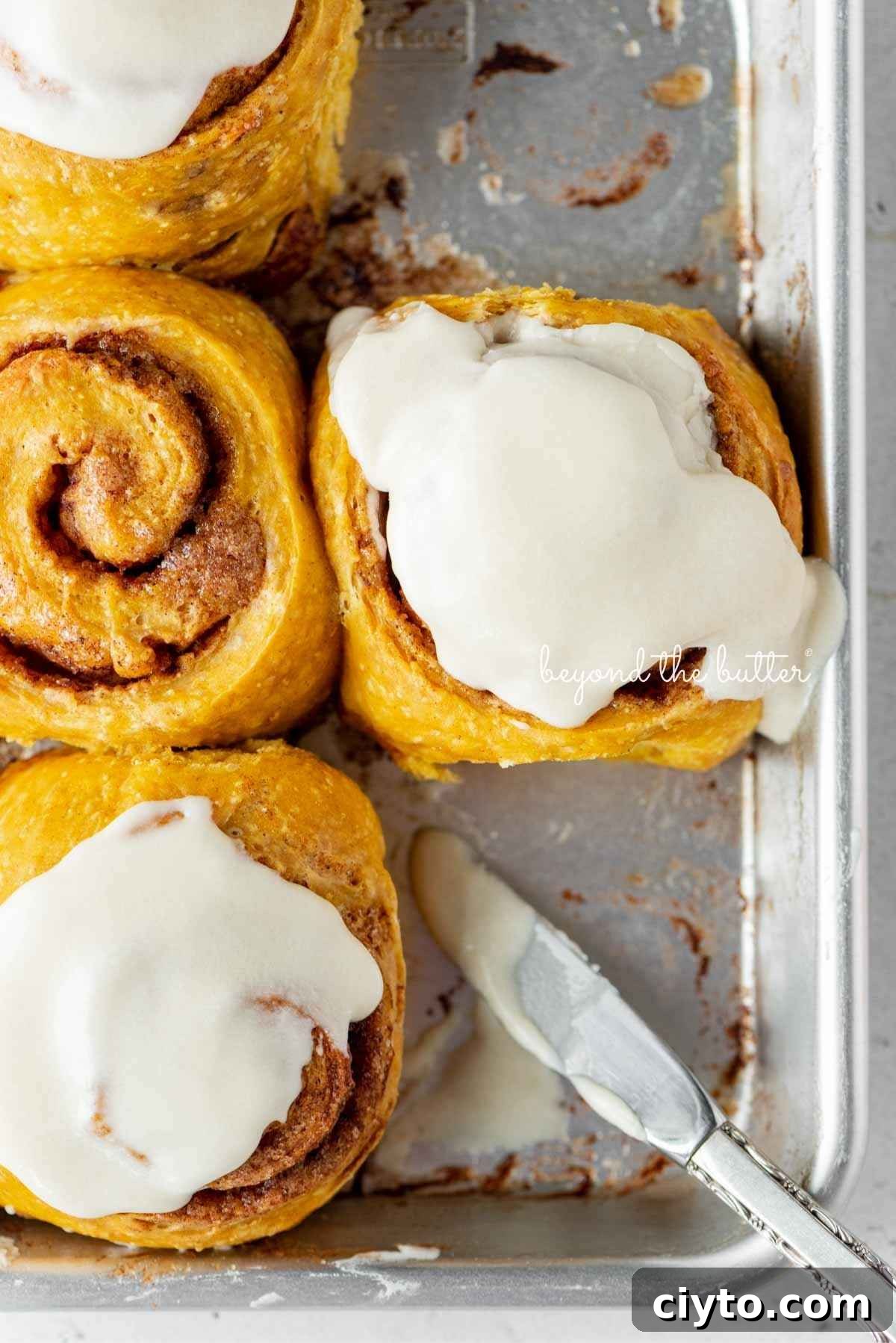 Four pumpkin cinnamon rolls on a small baking sheet with three topped with maple frosting.