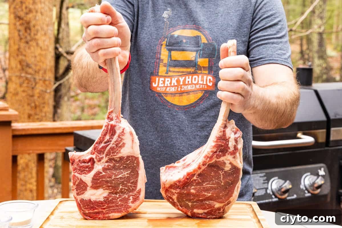Holding two large tomahawk ribeye steaks with a grill in background.