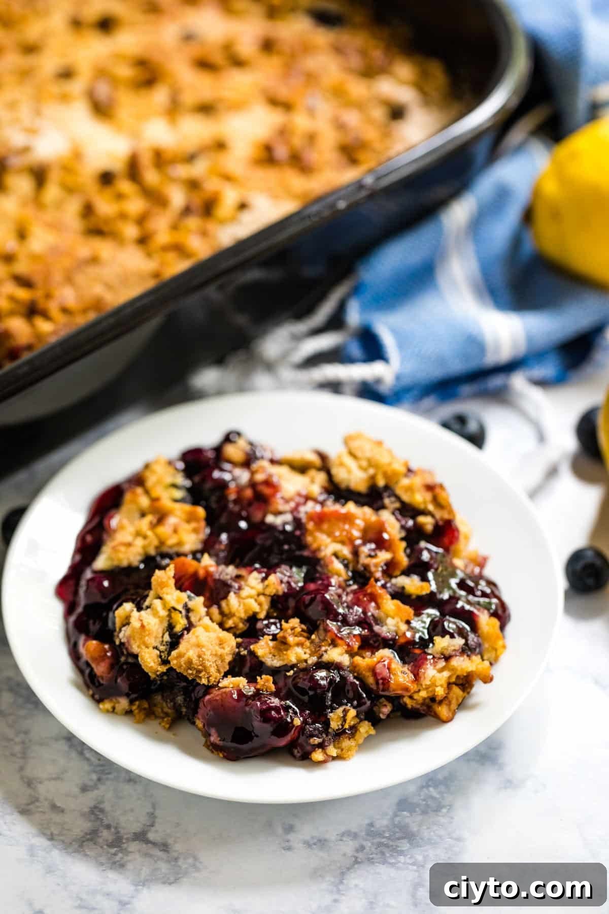 A white plate with lemon blueberry dump cake and the pan of dump cake behind it along with a blue and white striped towel and a lemon in the background.