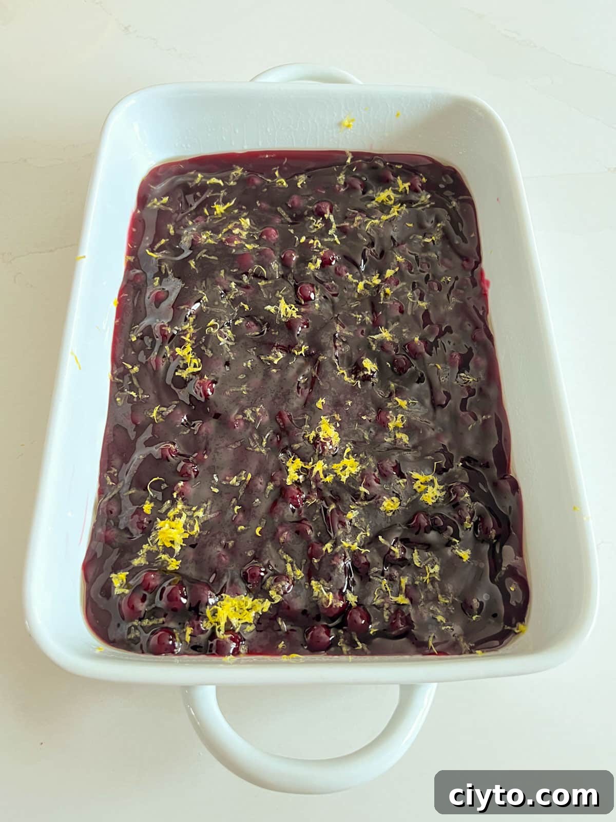 A white baking pan with blueberry pie filling spread in an even layer and topped with lemon zest.