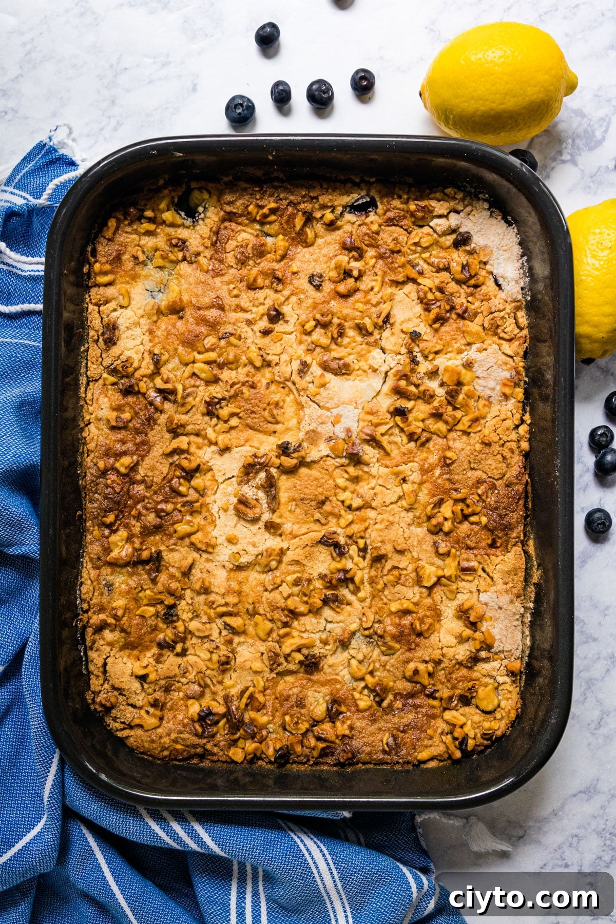 An overhead image featuring the top golden crust of the dump cake in a dark pan with a blue and white striped towel, blueberries and lemons around it.