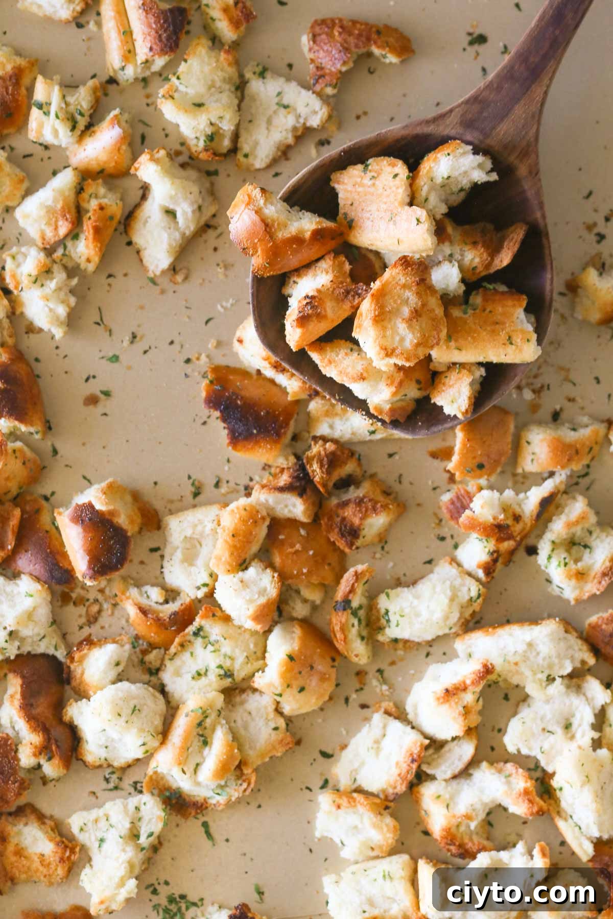 Overhead shot of a wooden spoon full of Rustic Buttery Garlic Croutons held over the baking sheet full of Rustic Buttery Garlic Croutons.