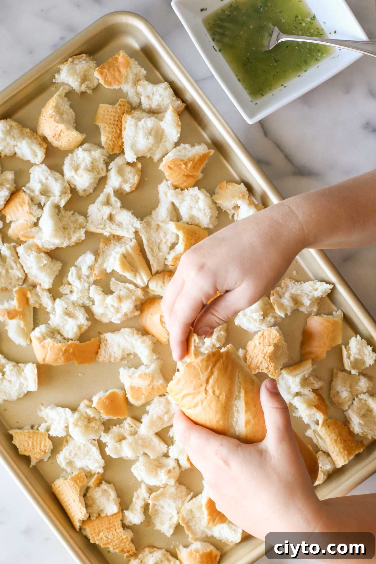 Overhead shot of a hand tearing the French bread into pieces over a baking sheet, with a square bowl of the butter mixture next to the baking sheet.