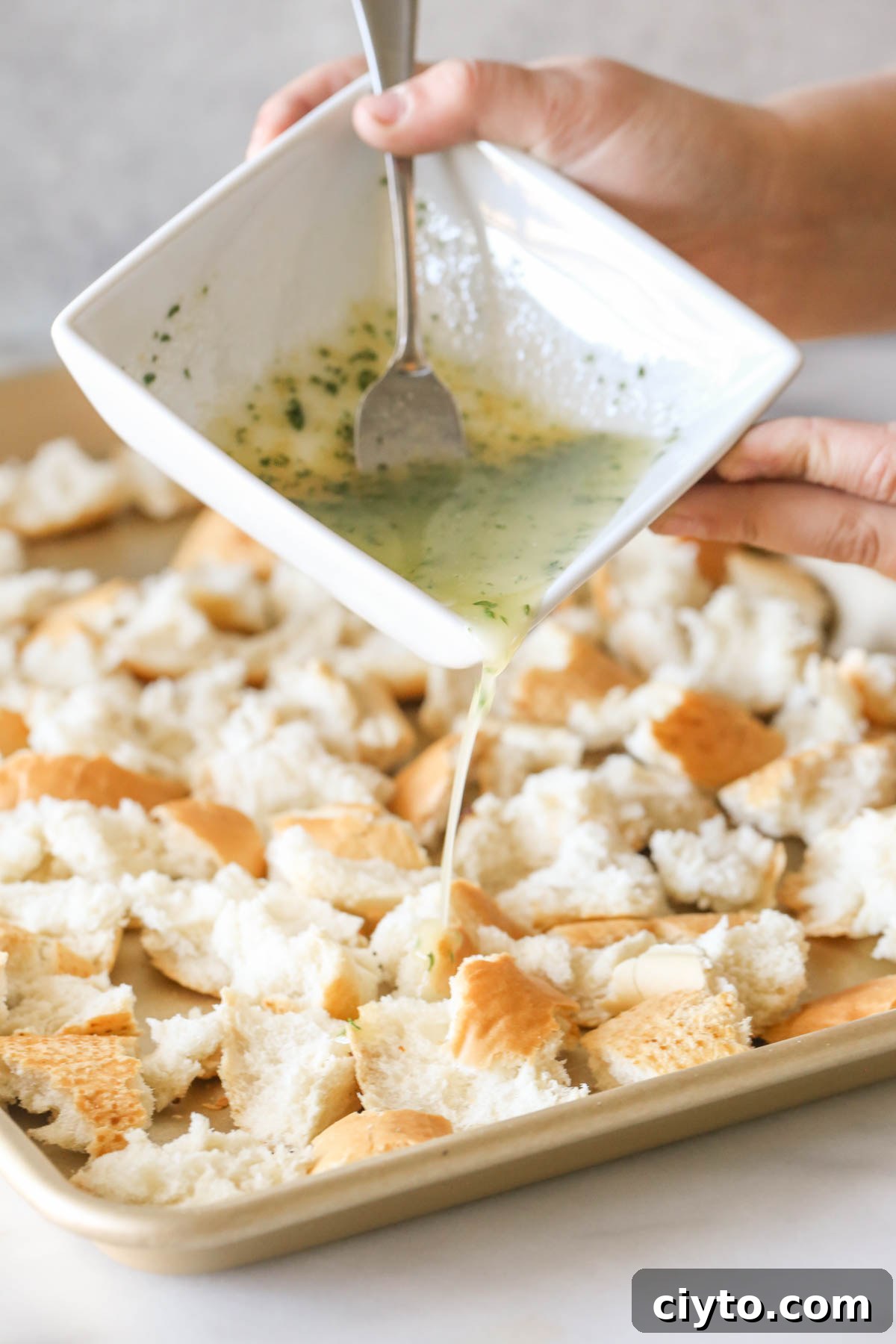 The butter mixture in a square bowl being poured over the French bread pieces on the baking sheet to make Rustic Buttery Garlic Croutons.