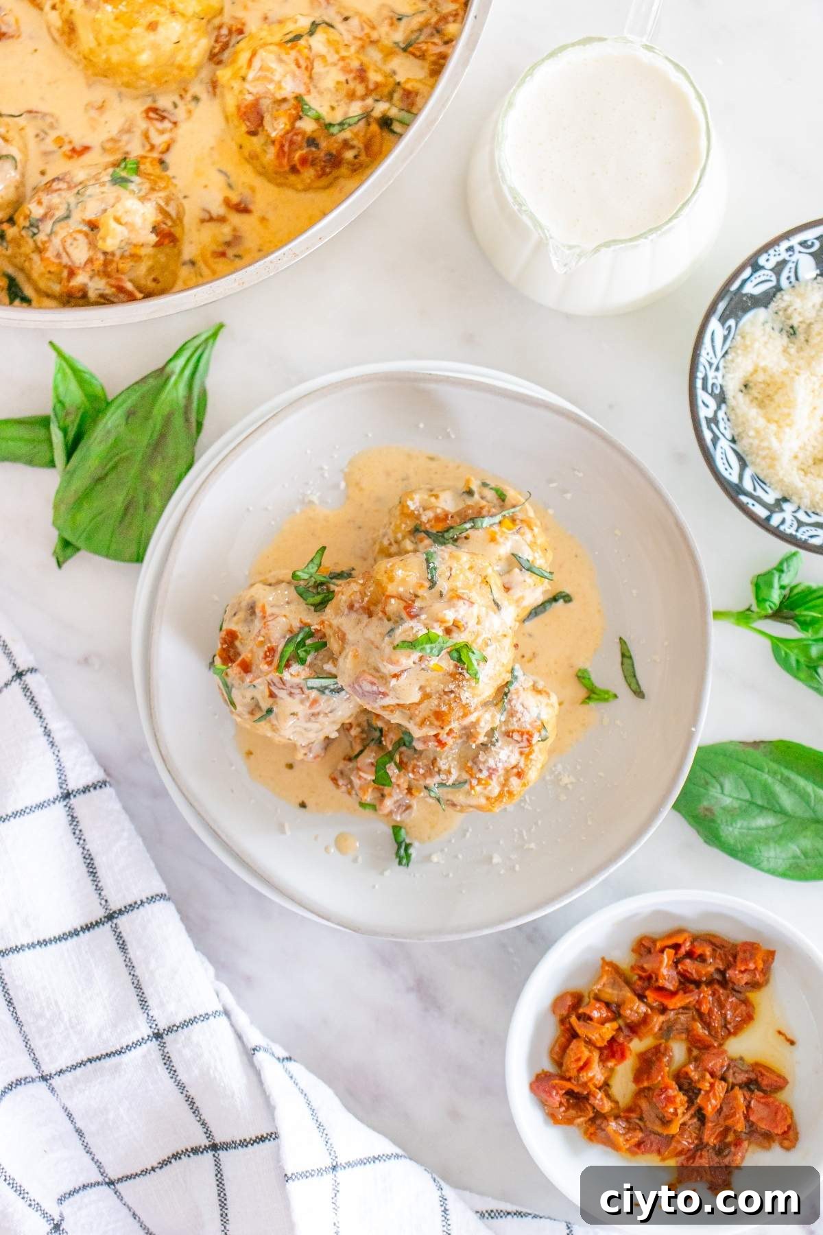 A plate of marry me chicken meatballs in creamy sauce garnished with herbs. Nearby are fresh basil leaves, a small bowl of Parmesan, a bowl of sun-dried tomatoes, and a striped cloth.
