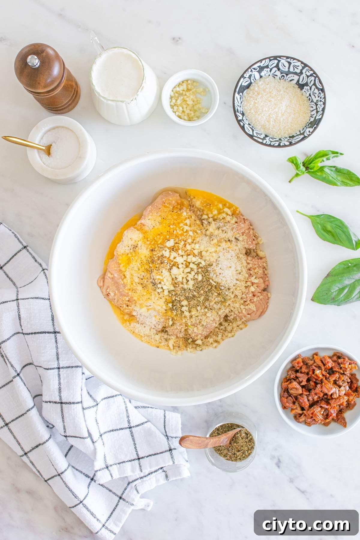 A bowl with mixed ingredients, featuring ground meat like in the famous marry me chicken meatballs, breadcrumbs, and seasonings. It’s surrounded by small bowls of spices, chopped vegetables, a pepper mill, and basil leaves on a marble surface.