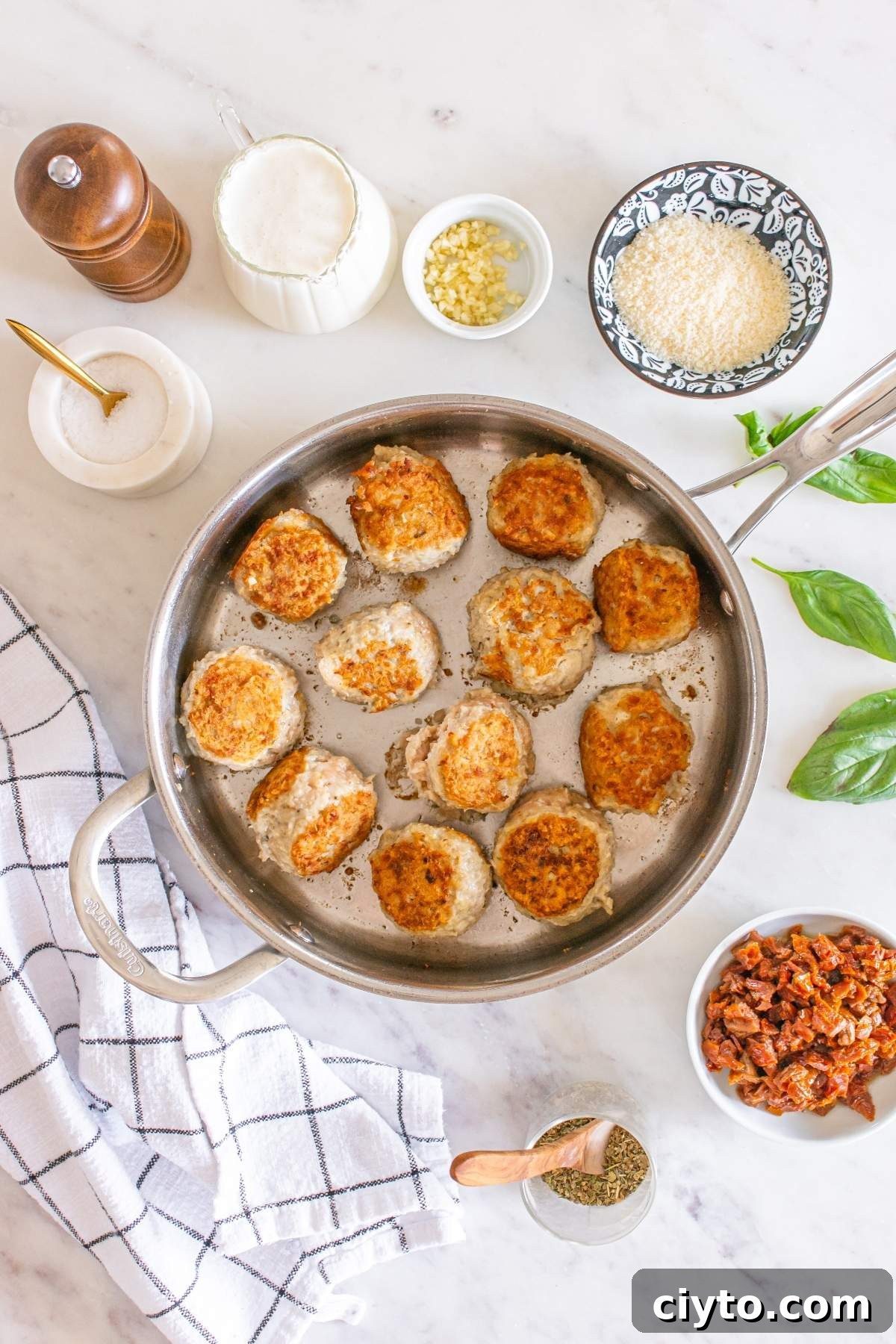 Fried meatballs in a pan are surrounded by a pepper mill, a cup of milk, minced garlic, breadcrumbs, fresh basil, sun-dried tomatoes, and salt on a marble countertop. These savory bites rival any marry me chicken meatballs recipe with their rich flavors.