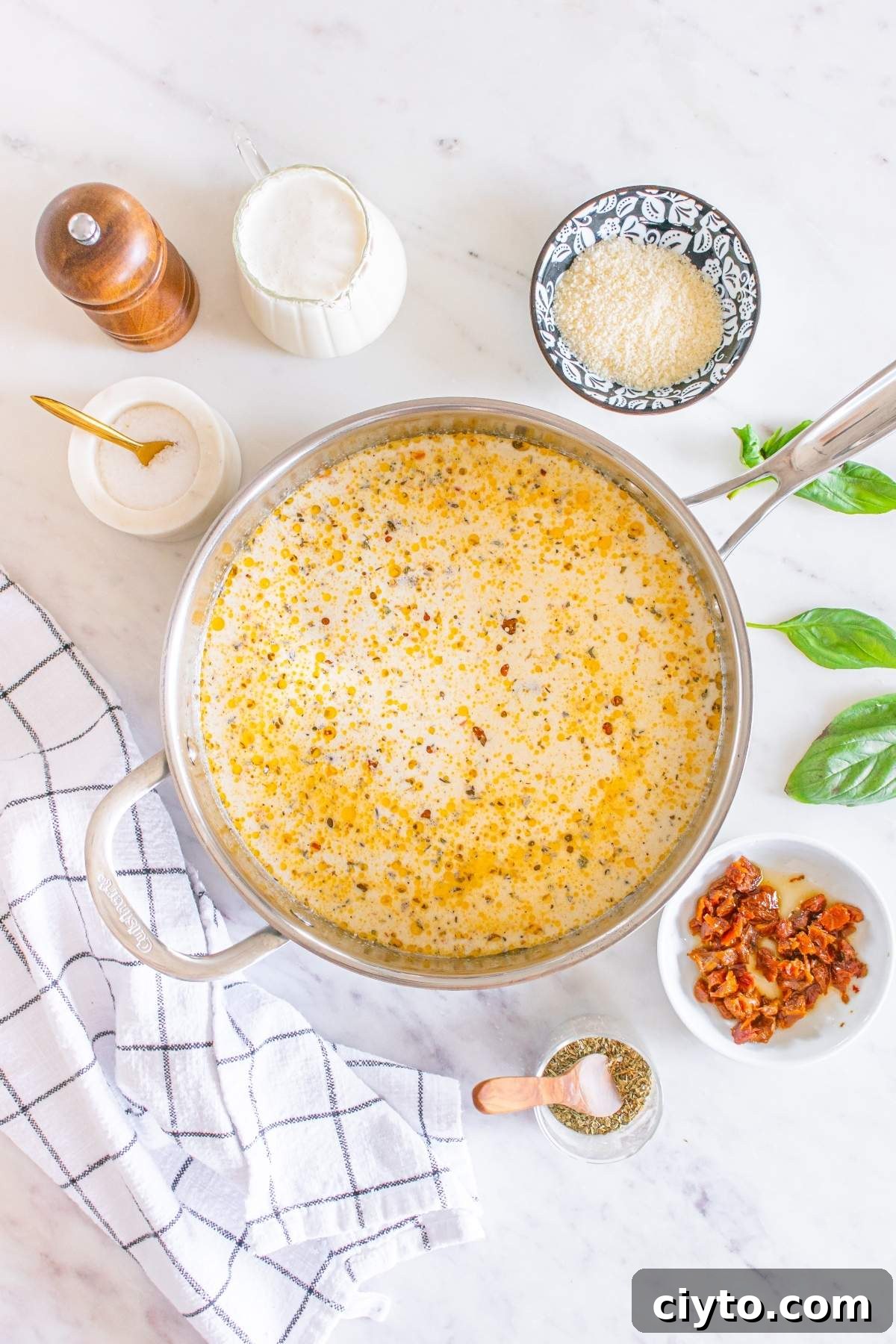 A pot of creamy soup sits on the countertop, its rich aroma mingling with the enticing scent of marry me chicken meatballs. Surrounding it are bowls of ingredients, a pepper grinder, a milk jug, fresh basil leaves, and a neatly folded cloth napkin.