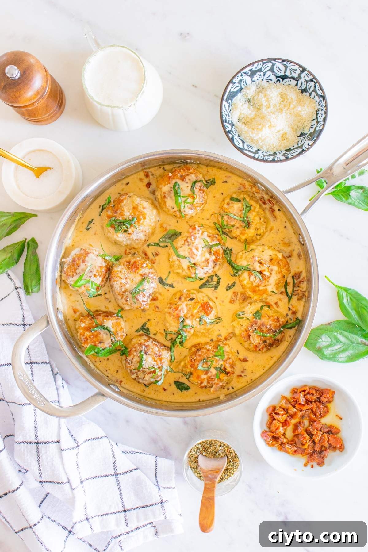 A skillet filled with "marry me" chicken meatballs in creamy sauce, garnished with herbs. Surrounded by bowls of grated cheese, crispy bacon bits, a pepper mill, and a cloth napkin.