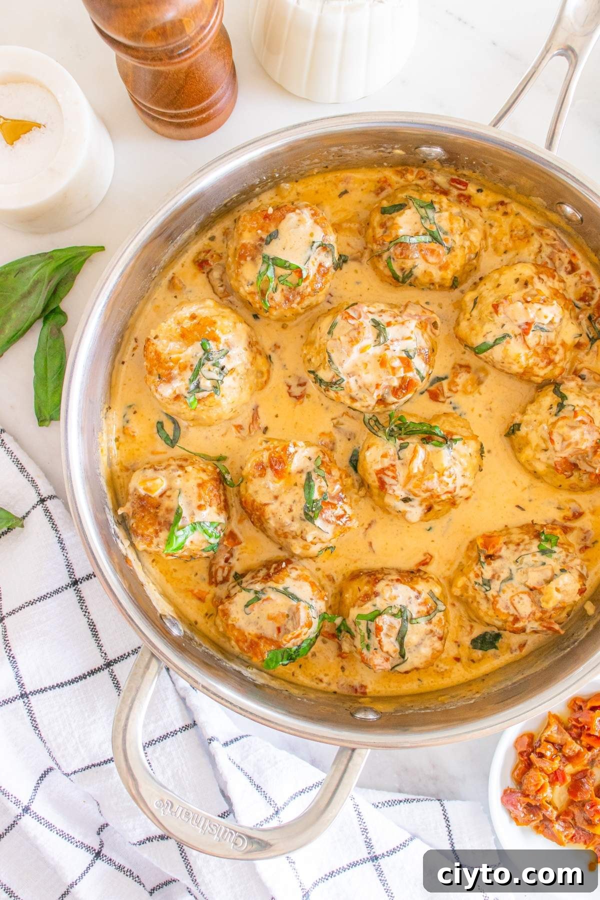 A skillet of creamy "marry me" chicken meatballs with herbs and sun-dried tomatoes, next to fresh basil leaves and a checkered cloth.