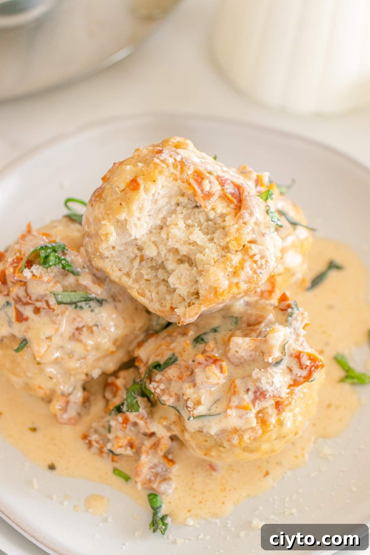 Three creamy "marry me" chicken meatballs topped with herbs sit on a white plate, accompanied by a rich sauce and some diced ingredients that enhance each savory bite.