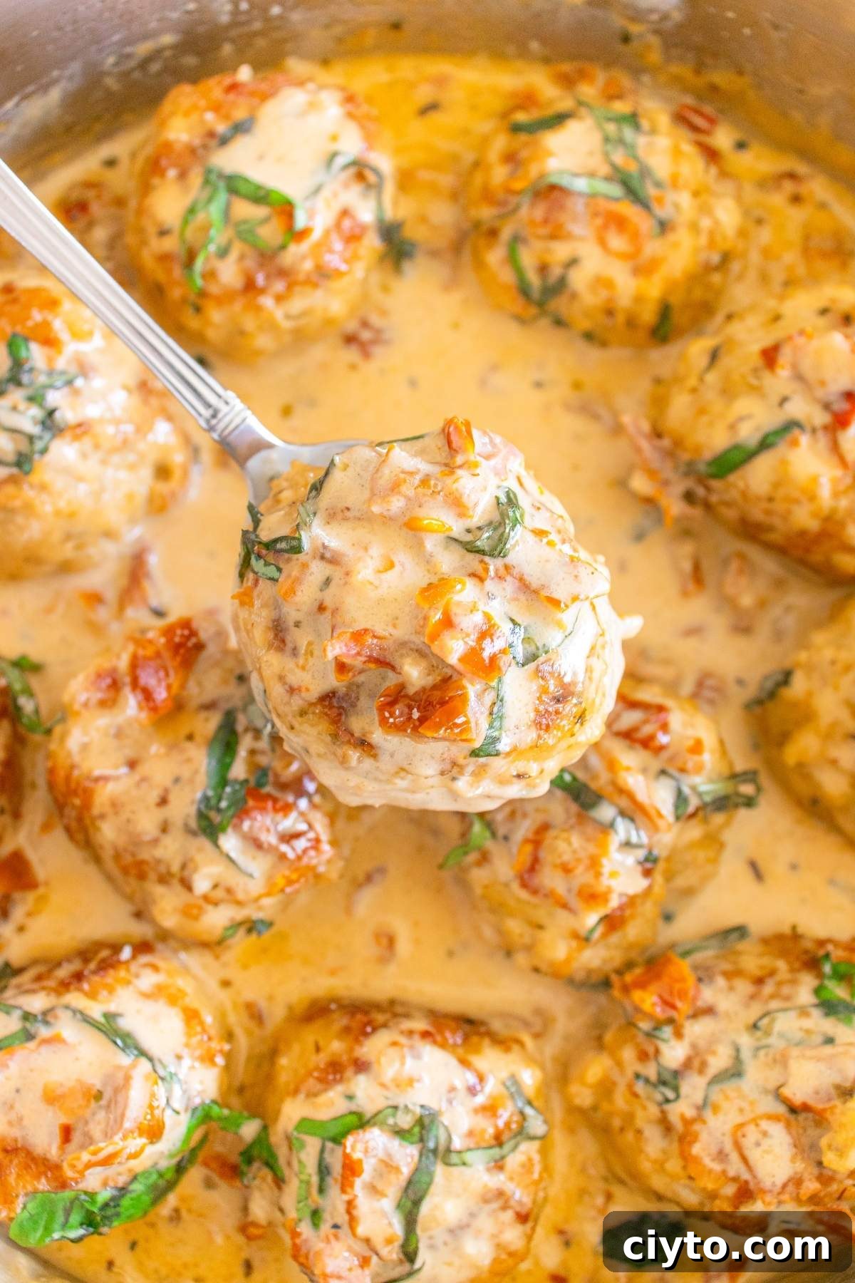 Close-up of creamy Tuscan marry me chicken meatballs in sauce, garnished with herbs and sun-dried tomatoes, with one meatball being lifted by a spoon.