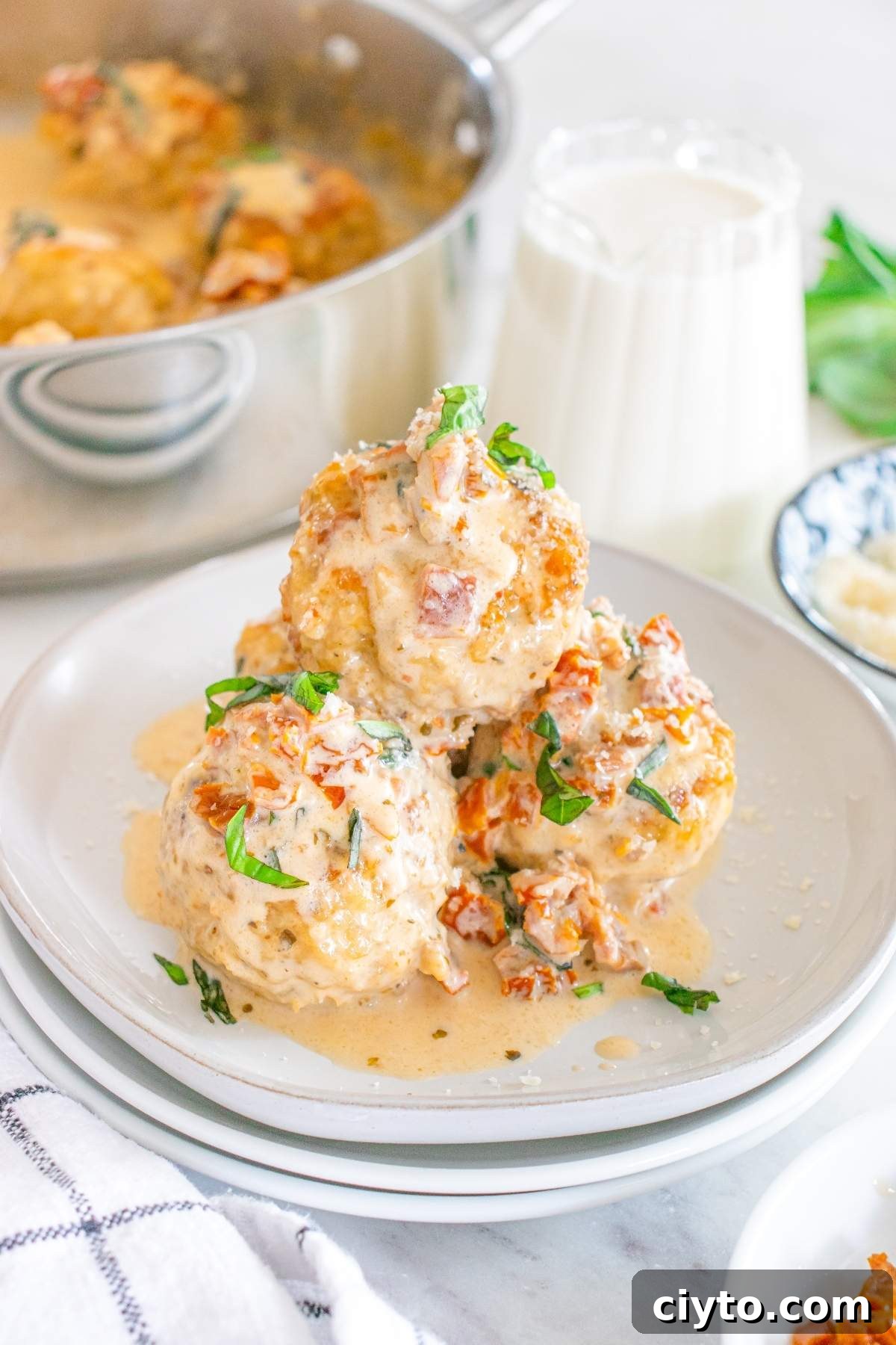 Three "marry me" chicken meatballs topped with creamy Tuscan sauce and herbs on a white plate. A pot and a glass of milk are in the background, adding a warm, inviting touch to this delightful meal.