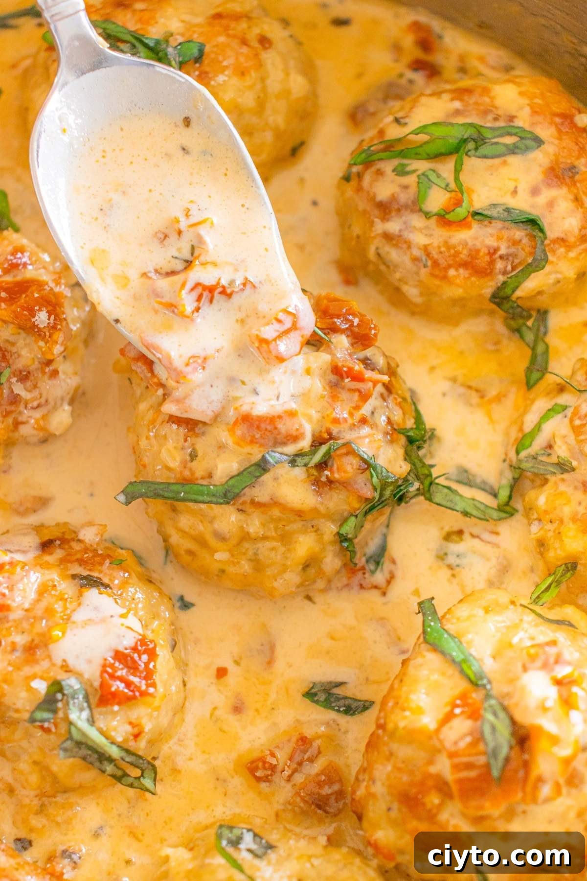 Marry Me chicken meatballs in a creamy sauce, garnished with chopped basil and sun-dried tomatoes, with a spoon drizzling sauce over one savory bite.