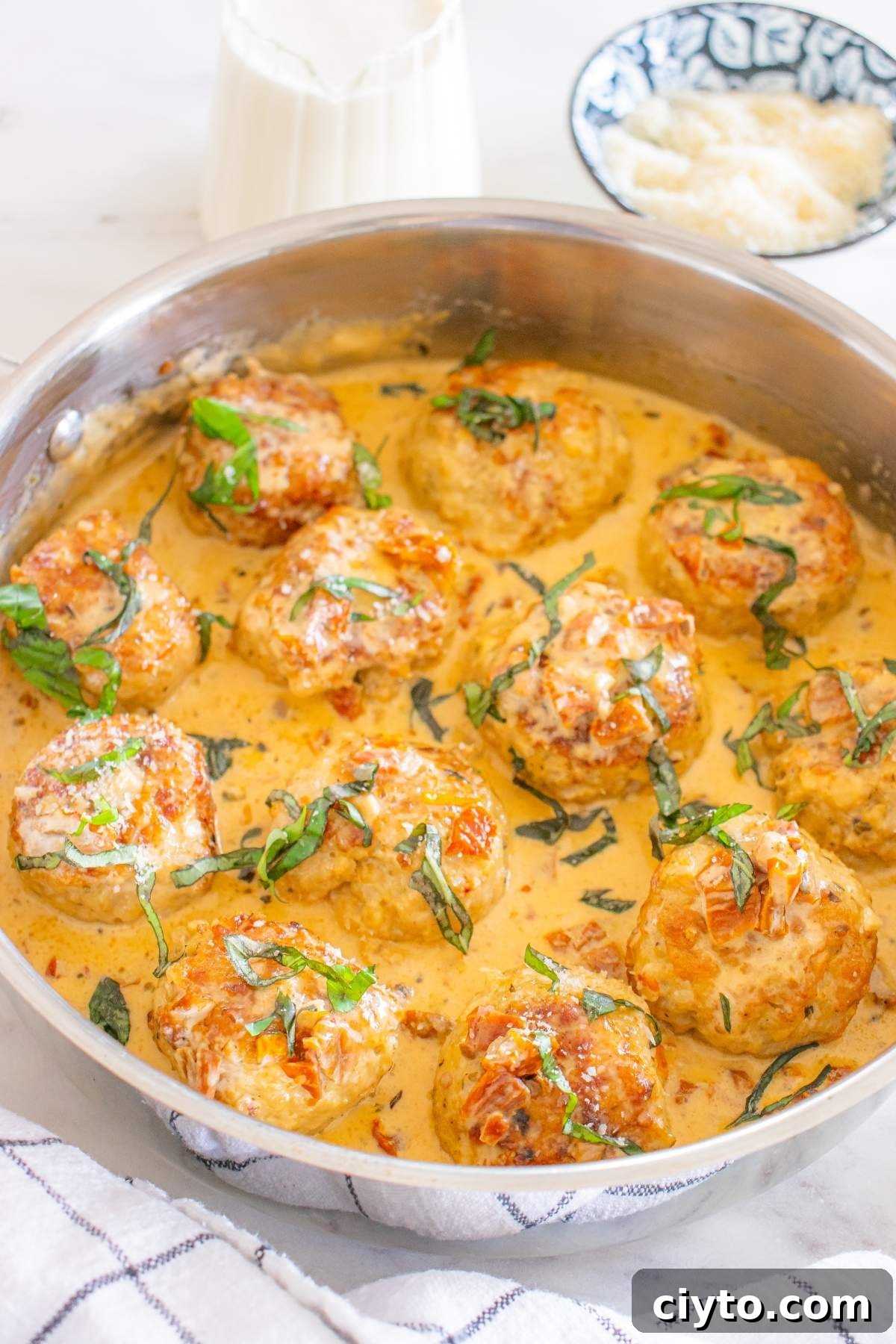Meatballs in a creamy Tuscan sauce (the beloved marry me chicken), garnished with sliced basil in a metal pan, with grated cheese and a milk pitcher nearby.