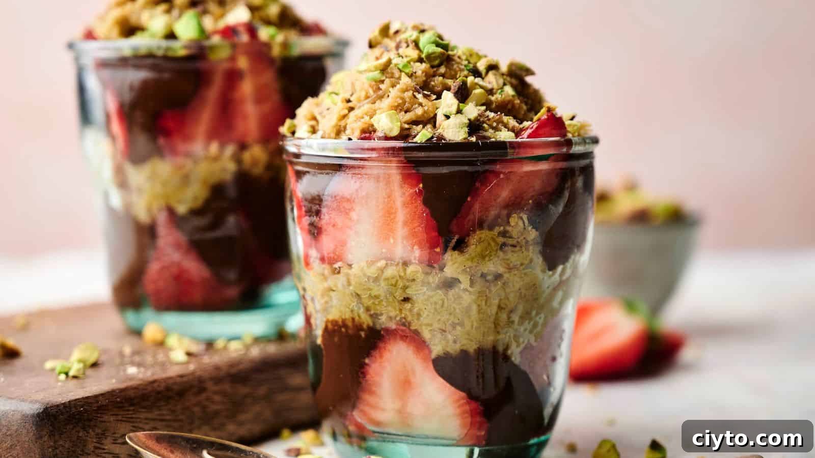 Two glass cups filled with layers of sliced strawberries, chocolate, granola, and chopped pistachios, with a blurred bowl of nuts in the background.