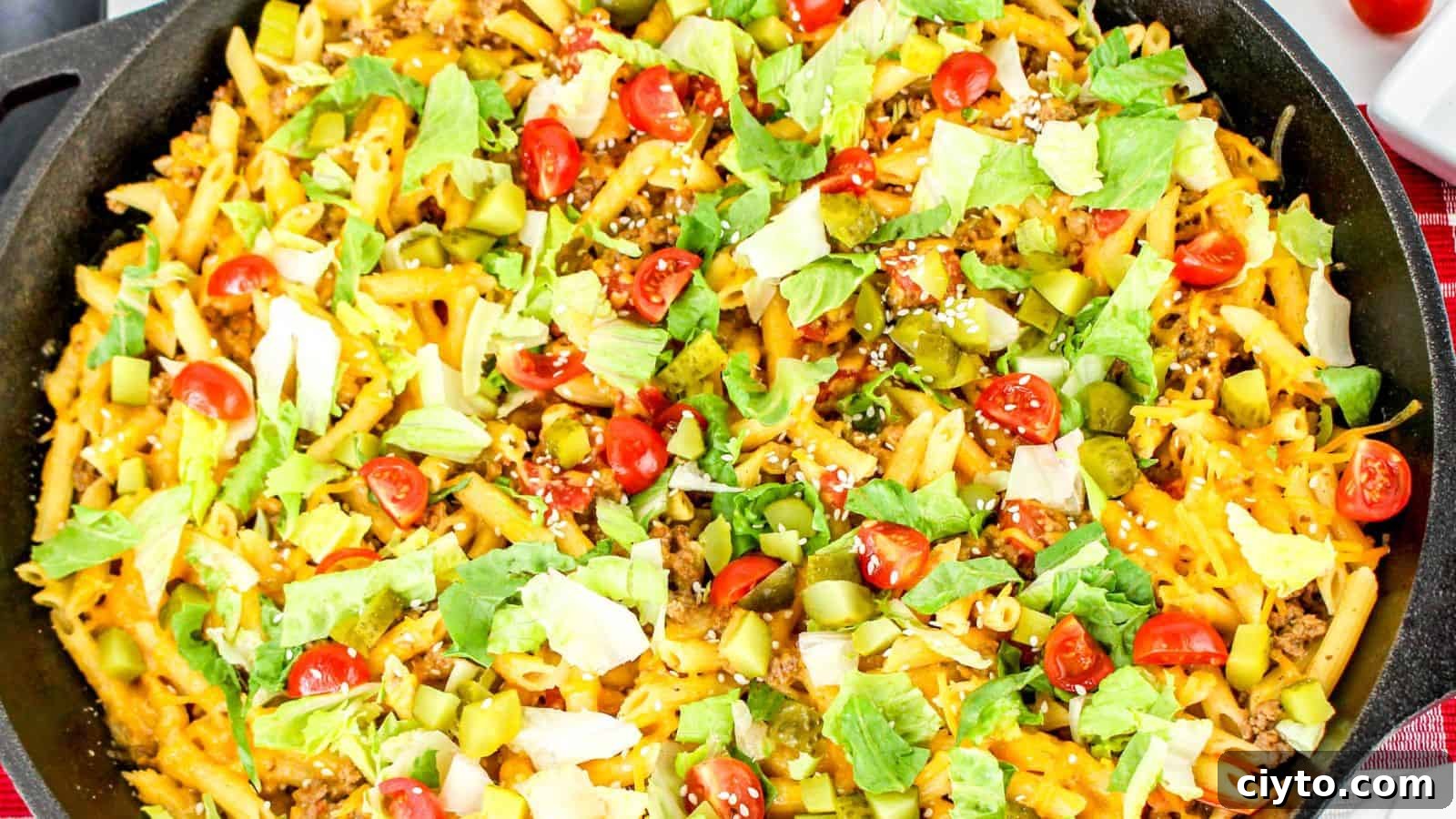 A skillet filled with penne pasta topped with shredded cheese, ground beef, diced tomatoes, lettuce, and pickles—a delicious take on cheeseburger pasta.
