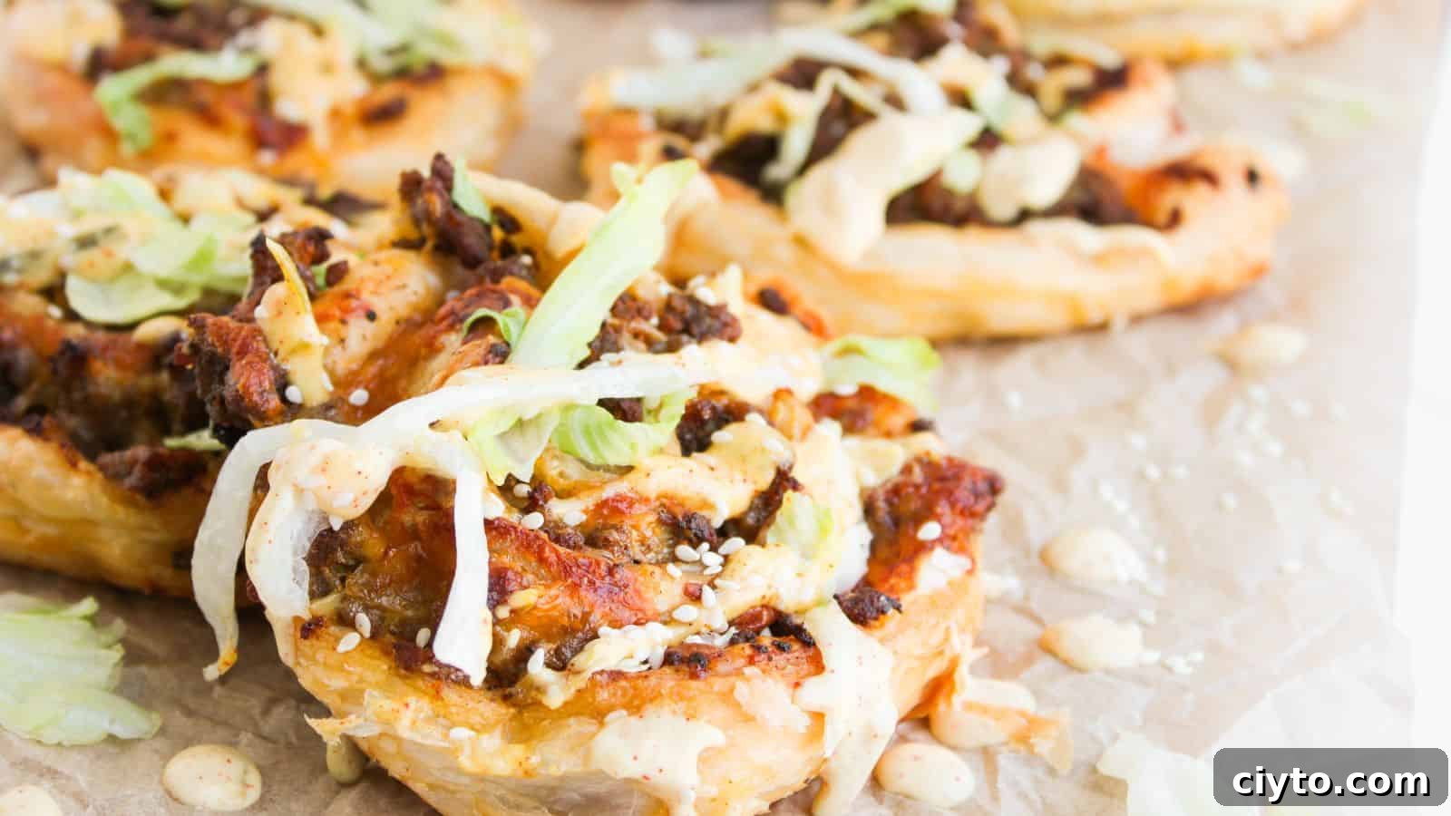 Close-up of puff pastry rounds topped with ground meat, shredded lettuce, sesame seeds, and a creamy sauce, arranged on parchment paper.