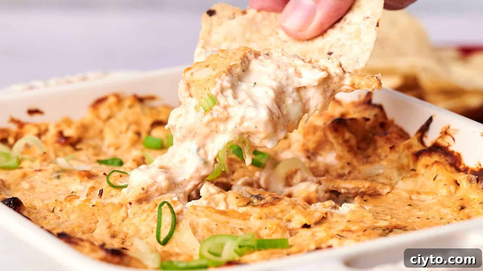 A hand dips a tortilla chip into a cheesy baked dish topped with sliced green onions.