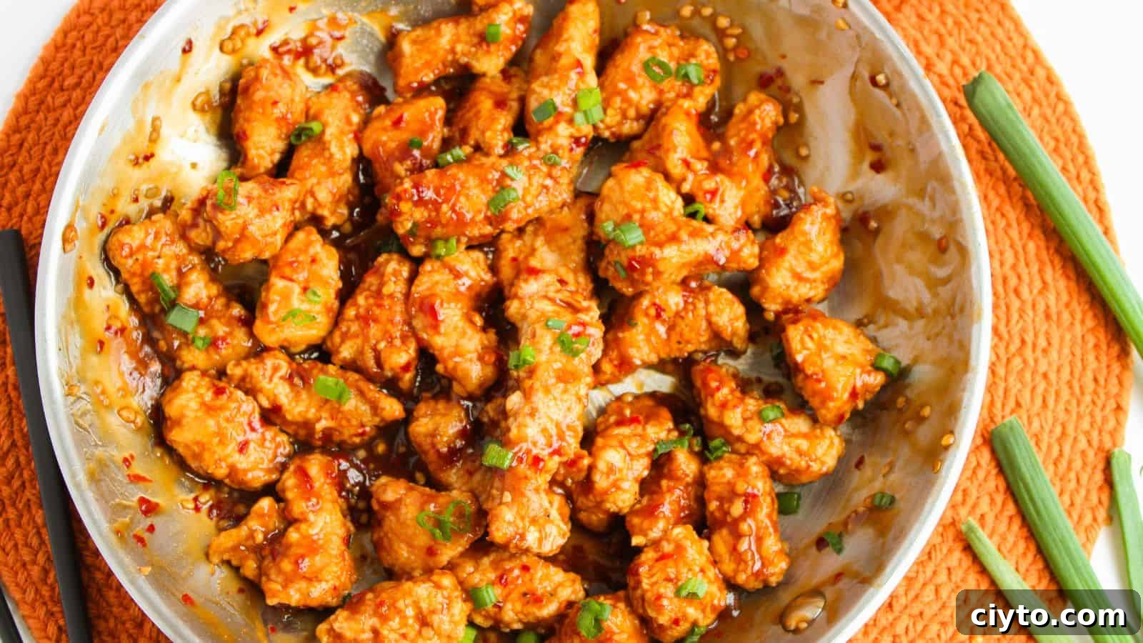 Stir-fried chicken pieces in a spicy sauce, garnished with chopped green onions, served in a round metal dish on an orange placemat.