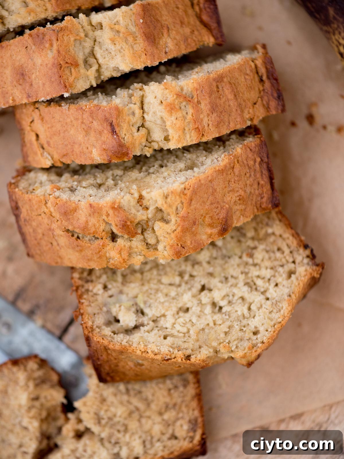 Sliced banana bread on a wooden surface, showing a moist and dense texture. The golden-brown crust suggests its freshly baked. A knife is partially visible at the edge.