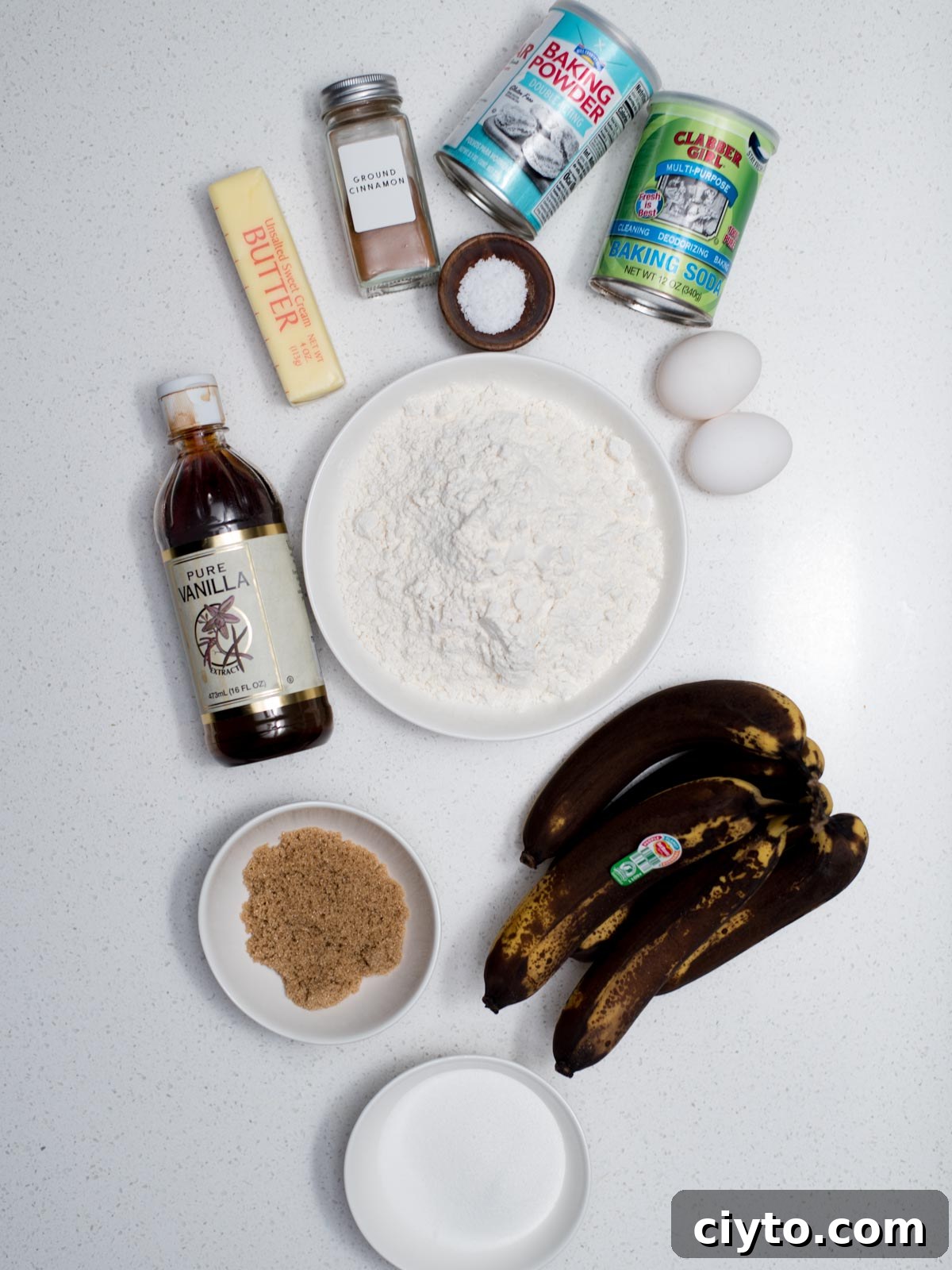 Ingredients for baking on a white surface: brown bananas, flour in a bowl, eggs, a stick of butter, vanilla extract, brown sugar, baking powder, baking soda, cinnamon, and salt.