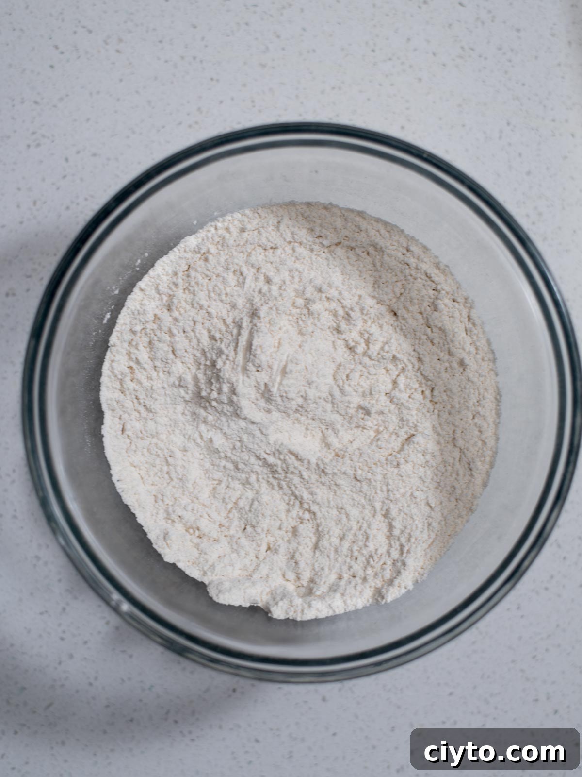 A glass bowl filled with white flour sits on a speckled white countertop. The flour appears finely milled and is slightly mounded in the center of the bowl.