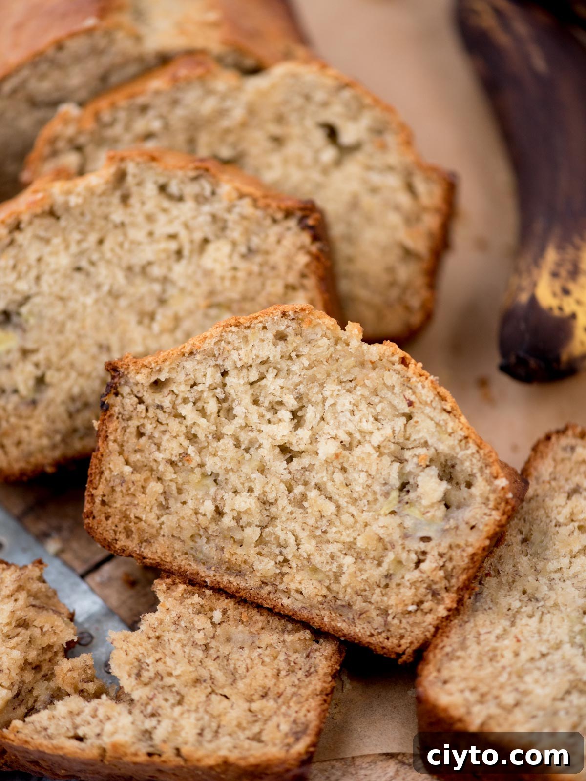 Slices of freshly baked banana bread on a wooden surface, showcasing its moist and fluffy texture. A partially visible ripe banana is in the background, adding context to the flavor of the bread.
