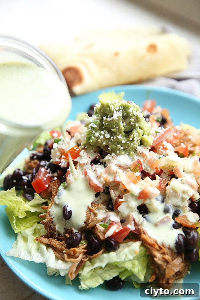 A light blue dinner plate with chopped lettuce topped with shredded chicken black beans, pico de Gallo, and guacamole being drizzled with dressing from a titled bottle on the left. Faded in the background is a folded tortilla.