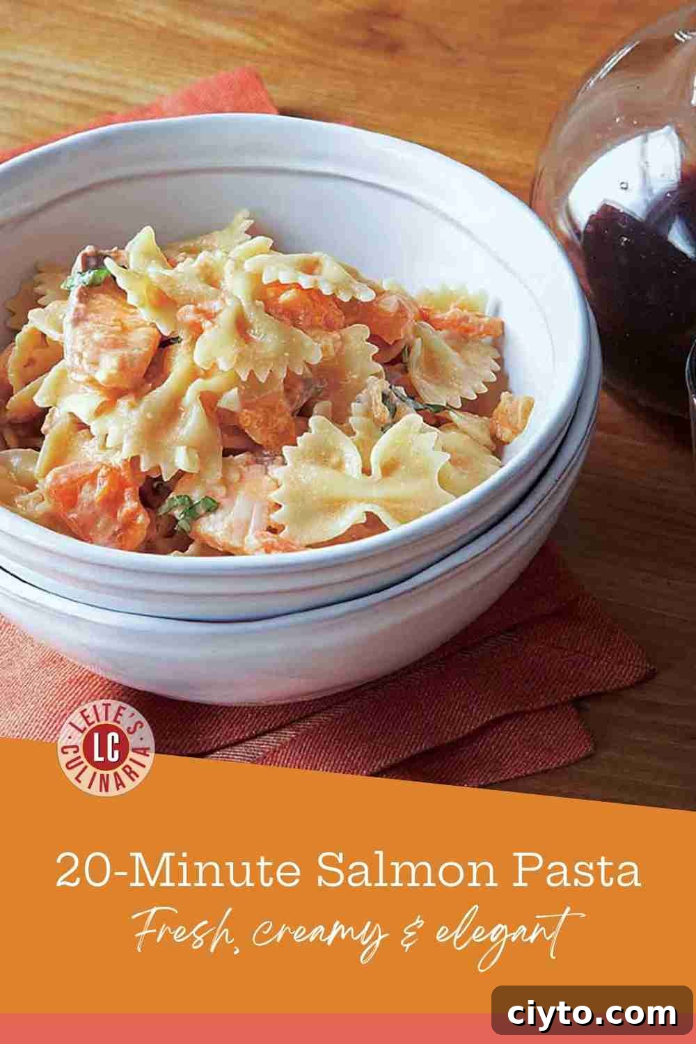 A white bowl of bow tie pasta with chunks of pink salmon in a light tomato cream sauce on a wooden table.