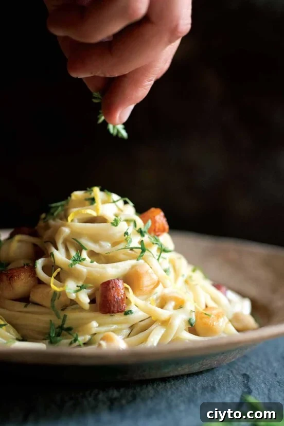 A tangle of fettuccine with scallops finished with lemon zest and chervil on a brown plate.