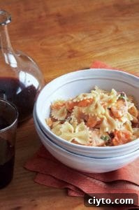 A white bowl filled with farfalle with fresh salmon is nested inside another white bowl with a carafe and glass of red wine in the background.
