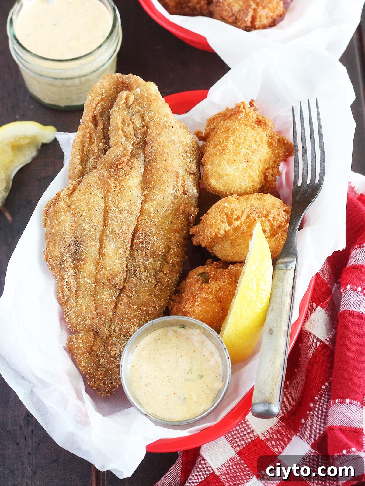 fried catfish fillet laying in a red basket with hushpuppies, remoulade sauce and lemon wedge