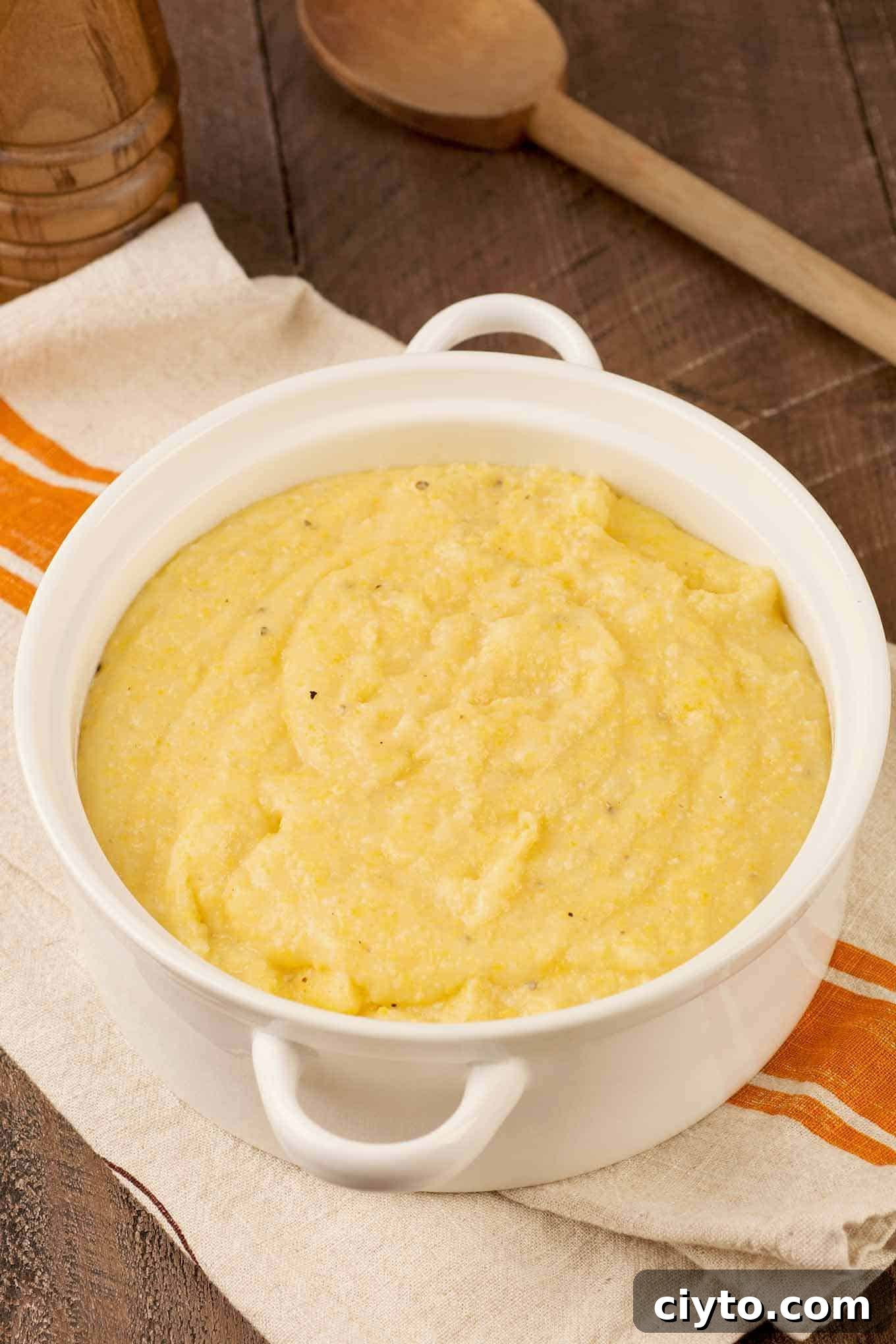 A round, white casserole of creamy polenta atop a striped tea towel with a wooden spoon to the rear.