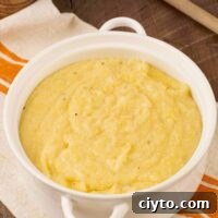 A round, white casserole of creamy polenta atop a striped tea towel with a wooden spoon to the rear.