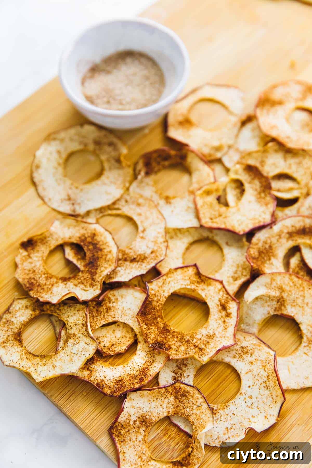 Easy homemade apple chips are crispy, chewy, rich, sweet, and covered in spicy cinnamon, making them the perfect fall snack. Overhead view of a bunch of apple chips on a cutting board, covered in cinnamon, next to a bowl of cinnamon sugar.