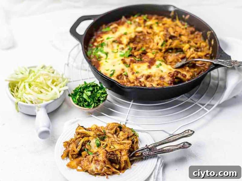 A cast iron skillet filled with baked pasta topped with melted cheese sits on a cooling rack, with a serving on a plate beside bowls of shredded cabbage and chopped herbs.