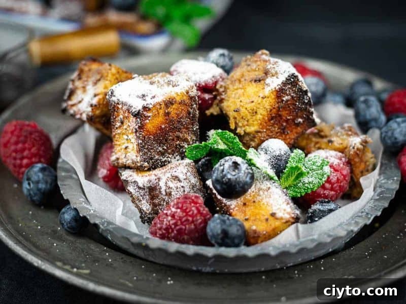 Cubes of French toast dusted with powdered sugar, served with fresh raspberries, blueberries, and a sprig of mint on a dark plate.