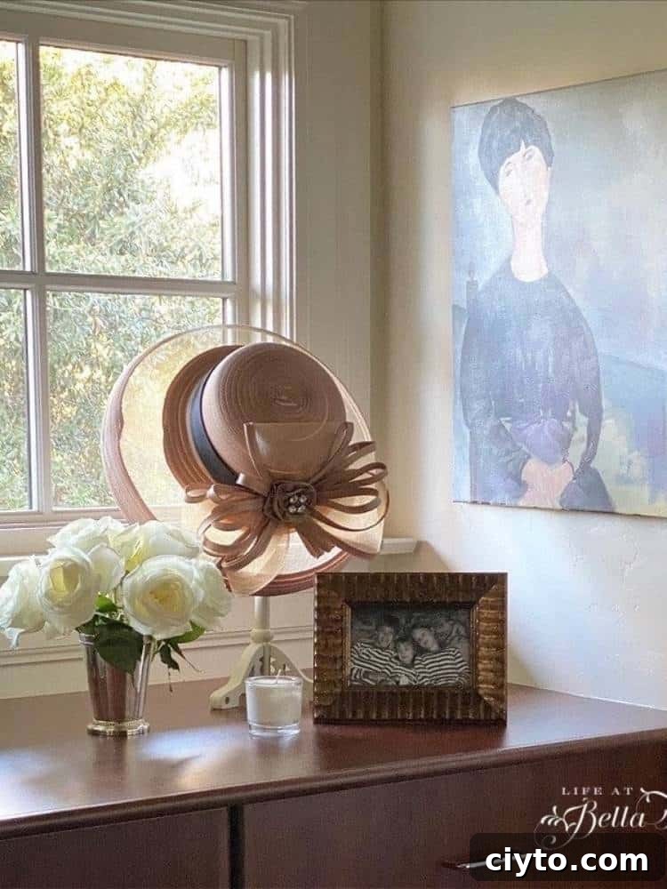 top of dresser in front of window with picture frame, vase of flowers, candle and a hat on stand displayed. print on wall to side above dresser top
