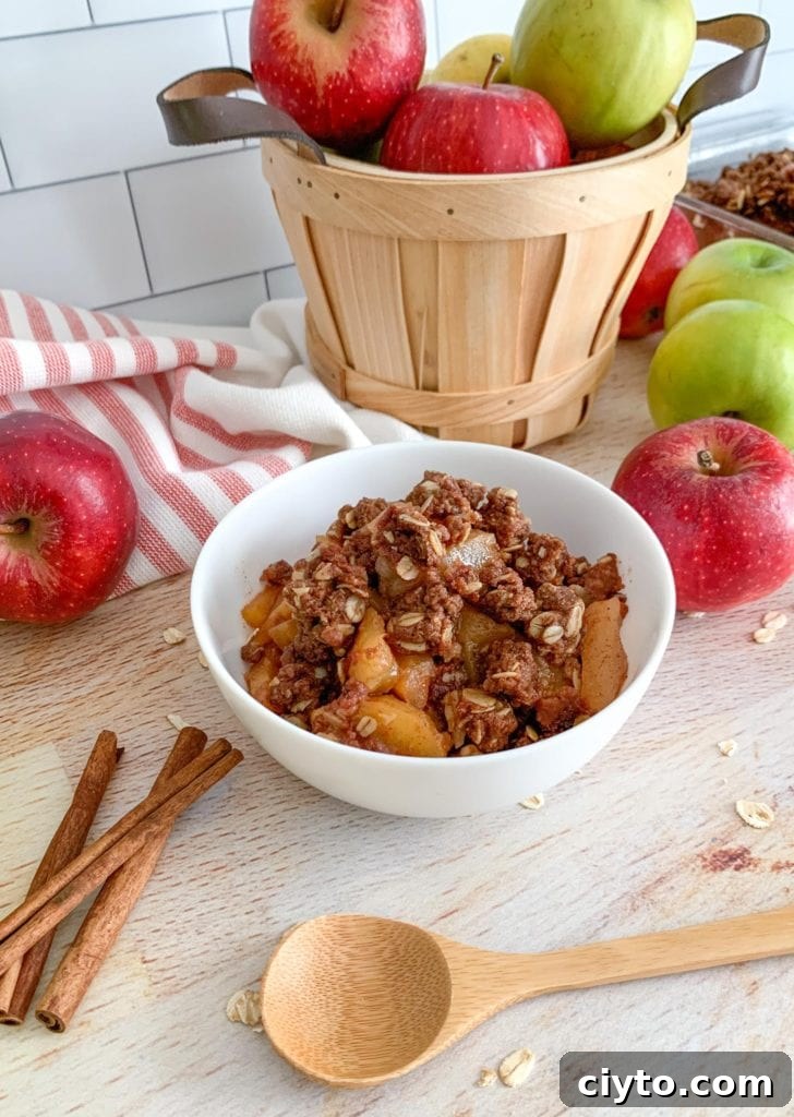 A bowl of apple crisp sitting on top of a butcher block table. Apples and cinnamon sticks are off to the side.